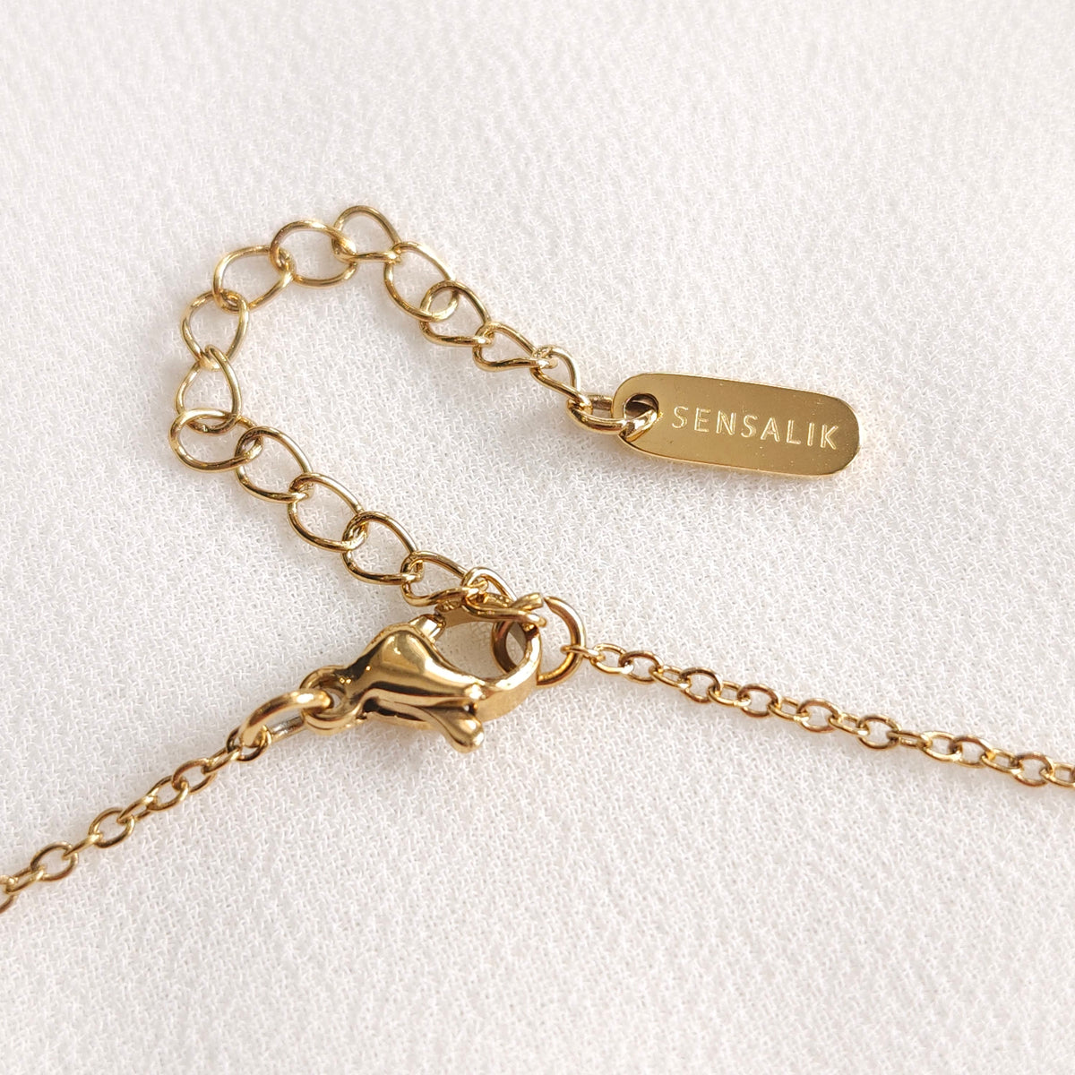 Close-up of the gold necklace’s lobster clasp, extension chain and engraved Sensalik tag.