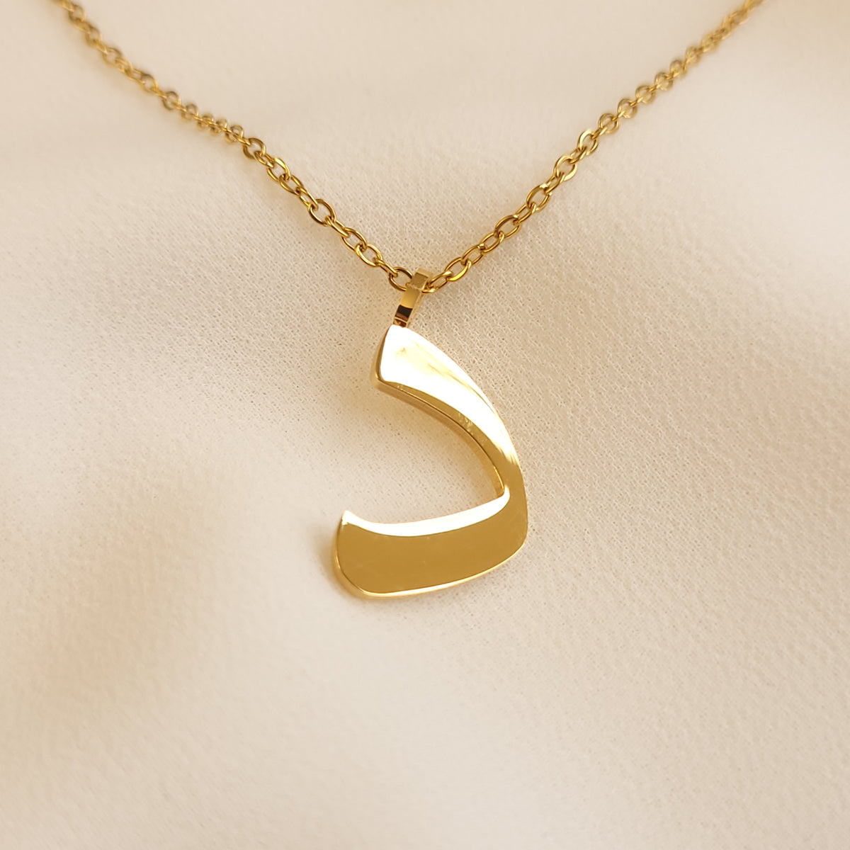 Gold-tone Arabic Initial Necklace with the letter Dal (د) on cream fabric.