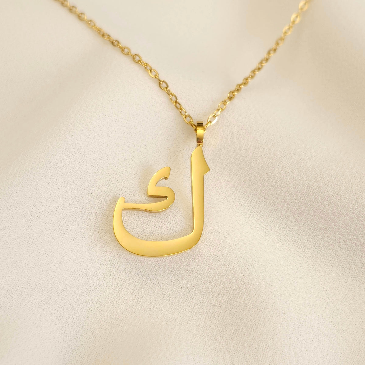 Gold-tone Arabic Initial Necklace with the letter Kaf (ك) on cream fabric.