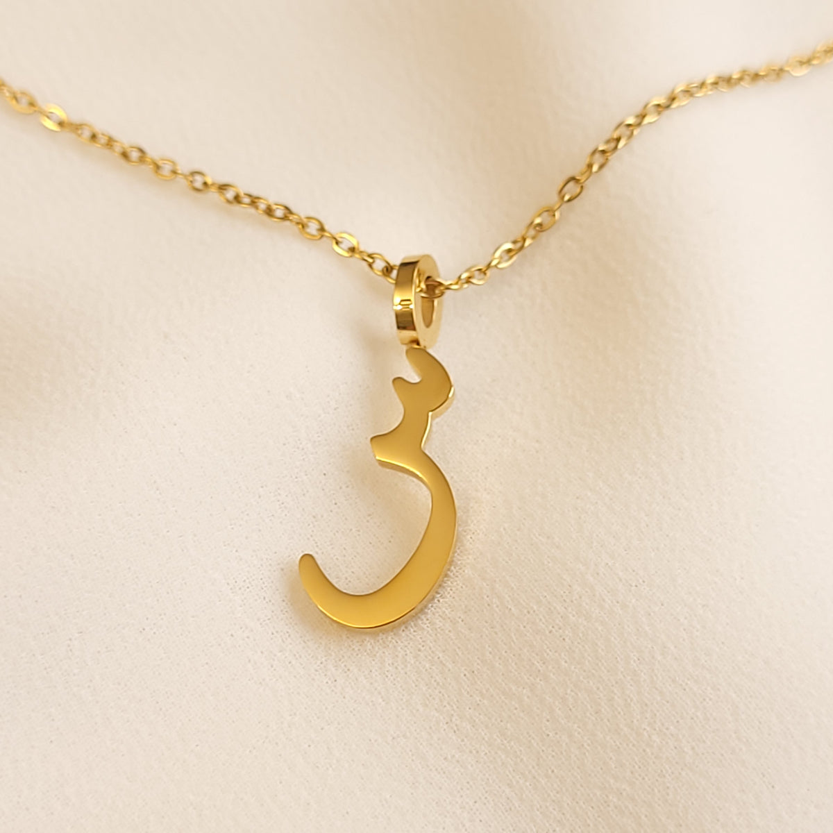 Gold-tone Arabic Initial Necklace with the letter Seen (س) on cream fabric.