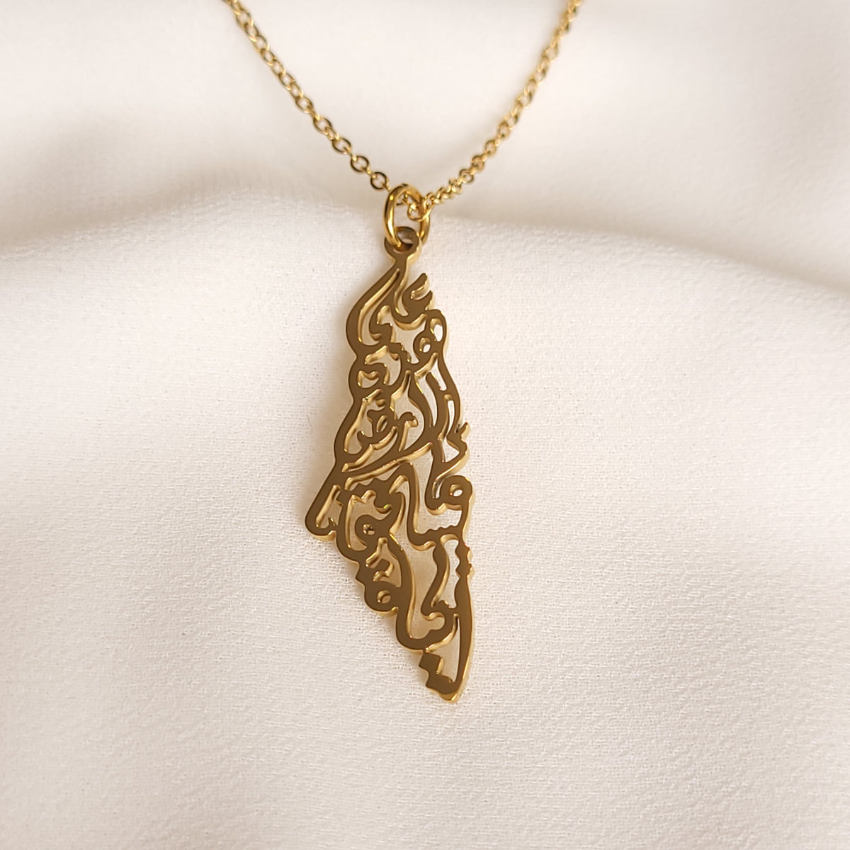Gold-tone Palestine Calligraphy Necklace showing the intricate map-shaped Arabic calligraphy pendant on cream fabric.