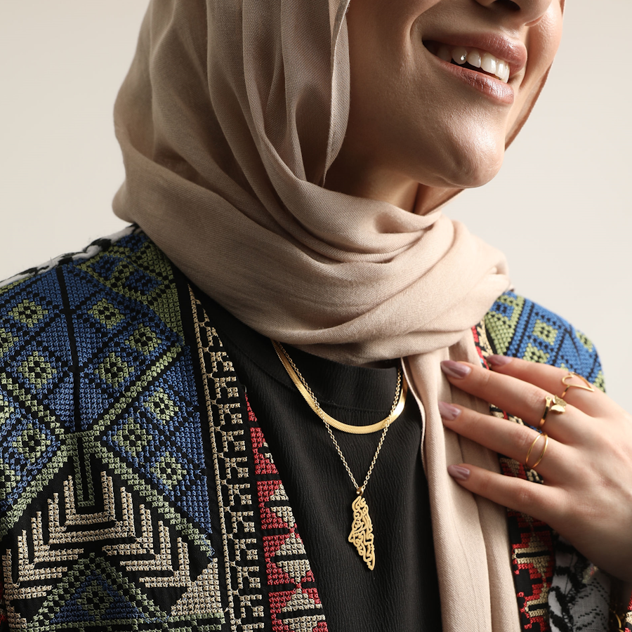 Model wearing the gold-tone Palestine Calligraphy Necklace layered with a shorter chain, a beige headscarf and an embroidered jacket.