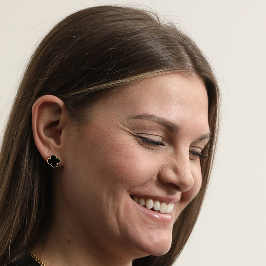 Close-up of a smiling model wearing a black Clover Stud Earring with a gold-tone border.
