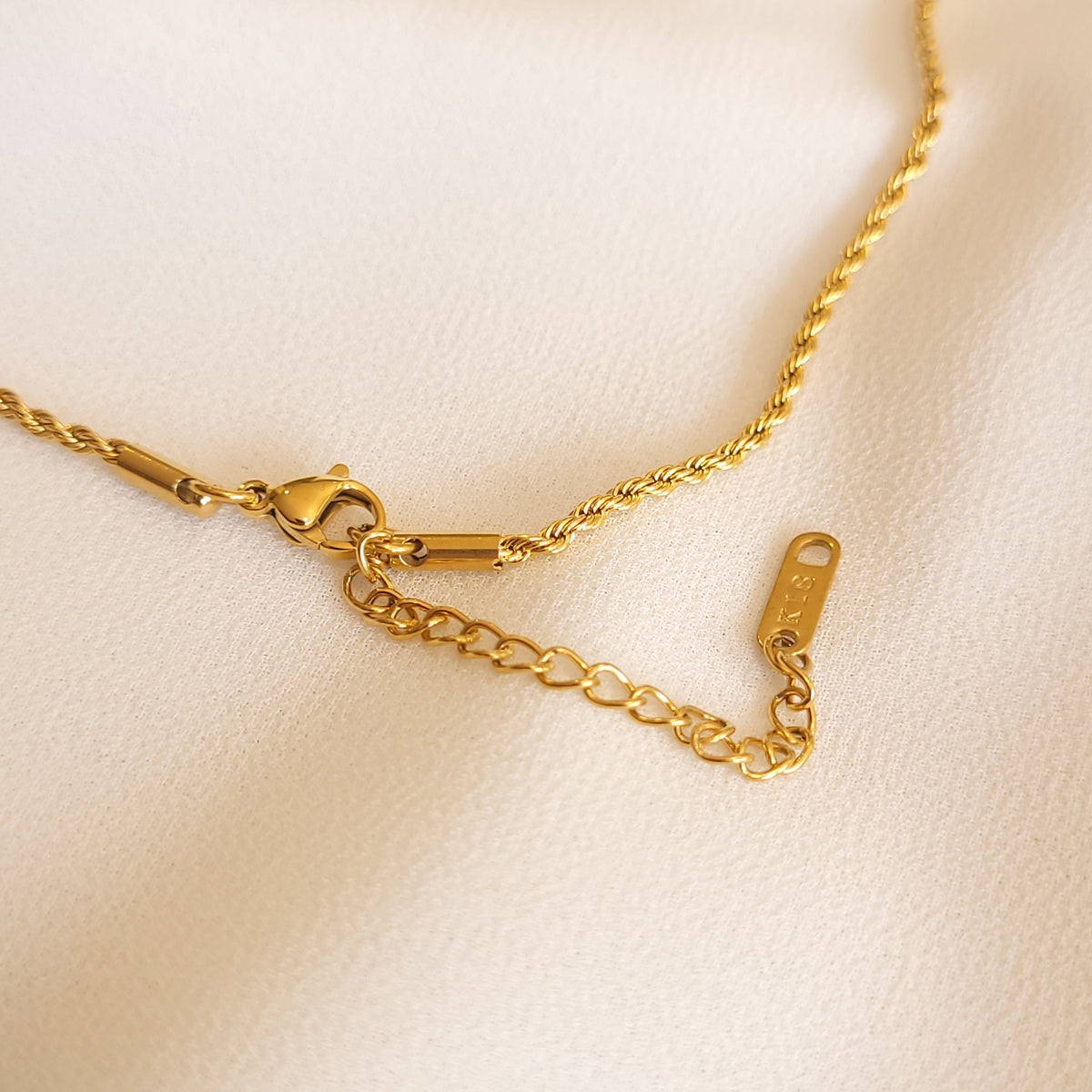 Close-up of the necklace’s gold-tone lobster clasp, extender chain and rectangular end tag on cream fabric.