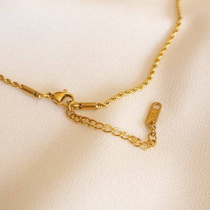 Close-up of the necklace’s gold-tone lobster clasp, extender chain and rectangular end tag on cream fabric.