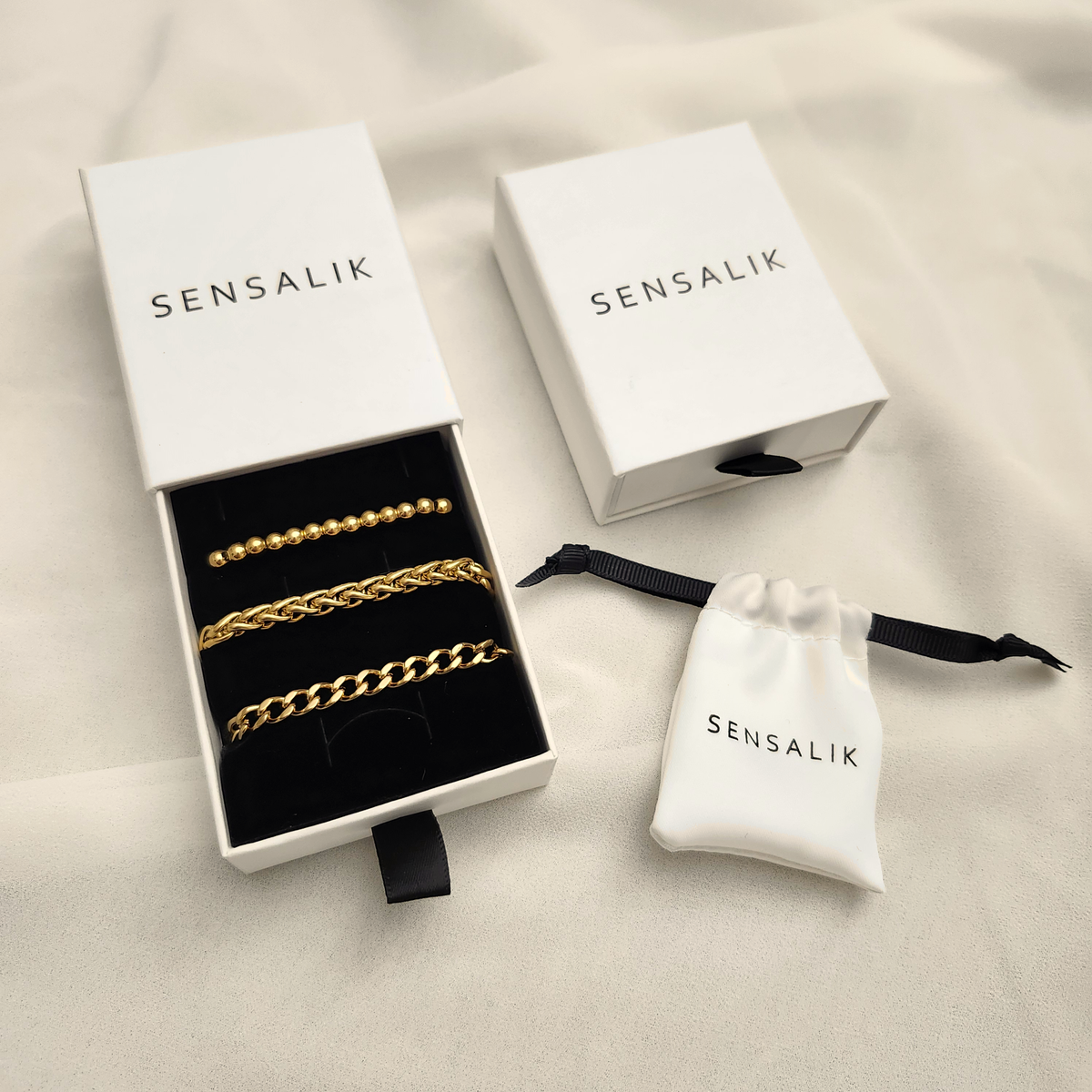 Gift Box opened to display gold-tone beaded and chain bracelets, beside a closed box and white drawstring jewelry pouch.