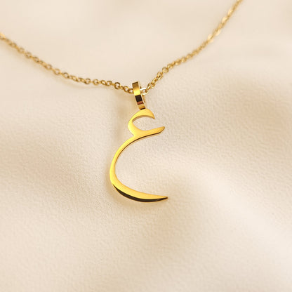 Gold-tone Arabic Initial Necklace with the letter Ain (ع) on cream fabric.
