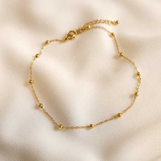 Gold-tone Satellite Beaded Anklet with evenly spaced round beads on a fine link chain, displayed on cream fabric.
