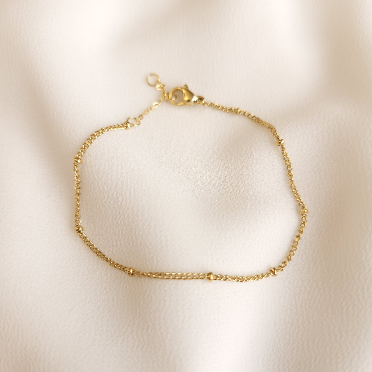 Gold-tone Satellite Beaded Bracelet with small spaced beads along a delicate chain on cream fabric.
