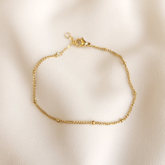 Gold-tone Satellite Beaded Bracelet with small spaced beads along a delicate chain on cream fabric.
