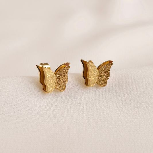 Gold-tone Sparkling Butterfly Stud Earrings with layered wings, textured fronts and polished edges on cream fabric.
