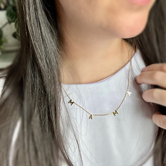 Mama Letter Necklace worn over a white top, showing the MAMA letter charms resting across the upper chest.

