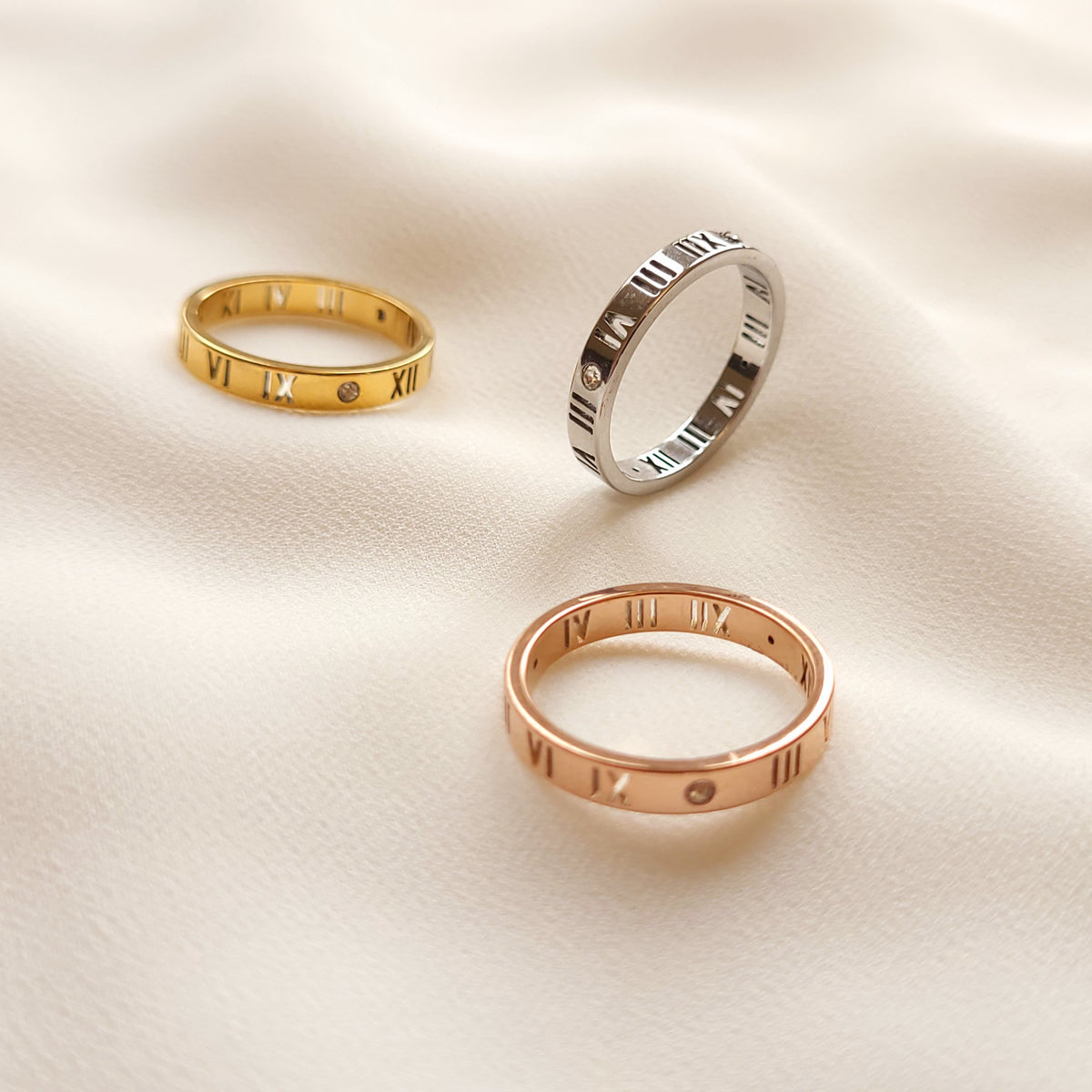 Roman Numeral Band Rings in gold, silver, and rose gold tones, showing their engraved numerals and clear stone accents.