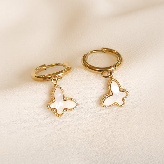 White Butterfly Drop Earrings with iridescent butterfly charms, beaded gold-tone borders and small hinged hoops on cream fabric.
